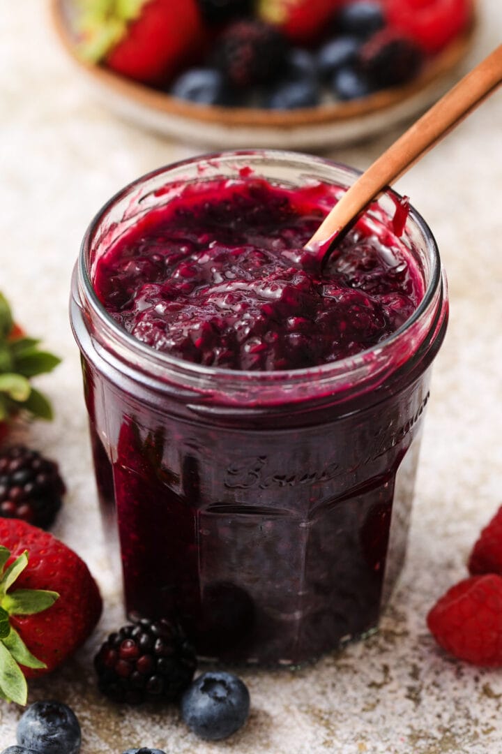 small jar of mixed berry jam, with a small speckled bowl of strawberries, raspberries, blueberries and blackberries off to the right hand upper corner.