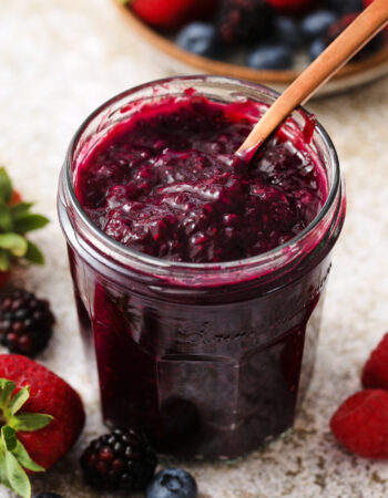 small jar of mixed berry jam, with a small speckled bowl of strawberries, raspberries, blueberries and blackberries off to the right hand upper corner.