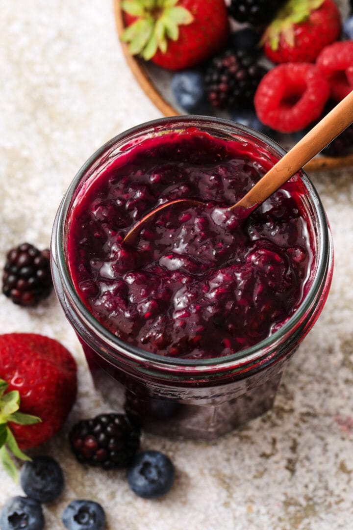 small jar of mixed berry jam, with a small speckled bowl of strawberries, raspberries, blueberries and blackberries off to the right hand upper corner.