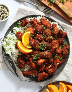 dark gray plate of orange chicken, piled on top of white rice and garnished with scallion and sesame seeds.