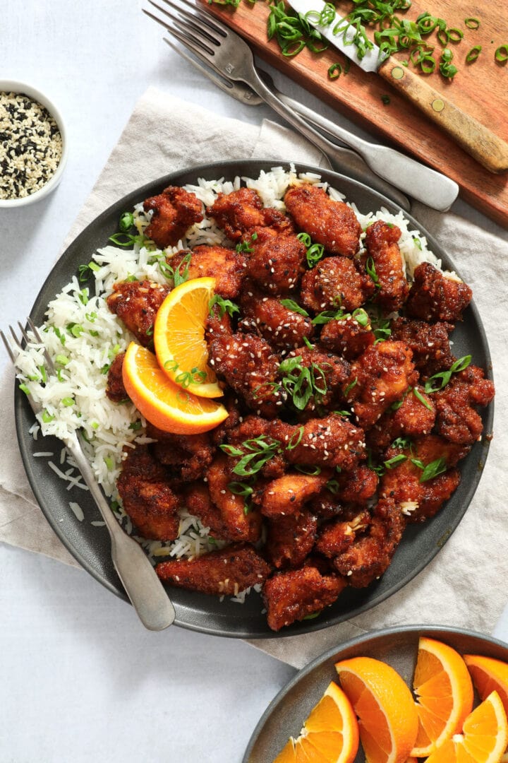 dark gray plate of orange chicken, piled on top of white rice and garnished with scallion and sesame seeds.