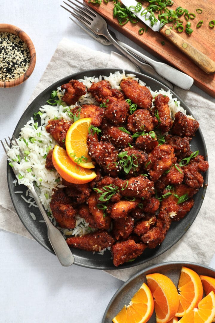 dark gray plate of orange chicken, piled on top of white rice and garnished with scallion and sesame seeds.