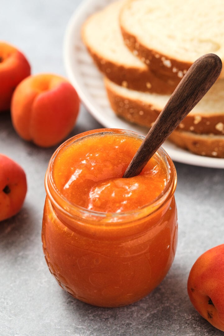 small jar of apricot jam with a tiny dark wooden spoon on top. There is a pile of toast and a few whole apricots in the background.