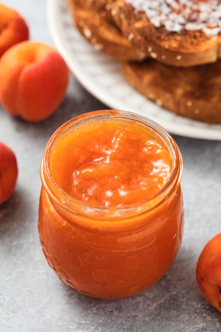 small jar of apricot jam with a tiny dark wooden spoon on top. There is a pile of toast and a few whole apricots in the background.