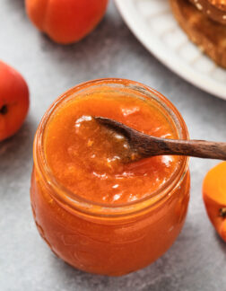 small jar of apricot jam with a tiny dark wooden spoon on top. There is a pile of toast and a few whole apricots in the background.