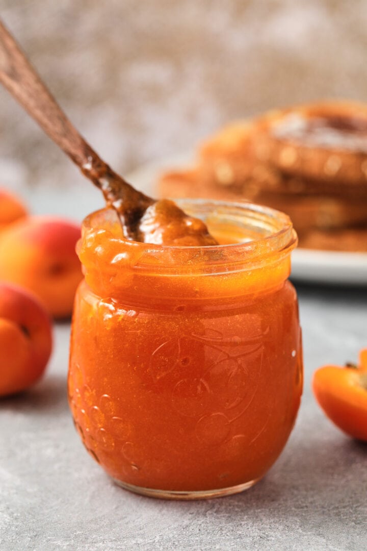 small jar of apricot jam with a tiny dark wooden spoon on top. There is a pile of toast and a few whole apricots in the background.