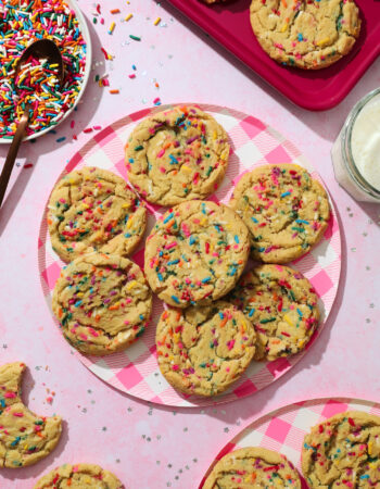 pink gingham print plate with several rainbow sprinkle cookies and a glass of milk off to the right hand side of the photo.