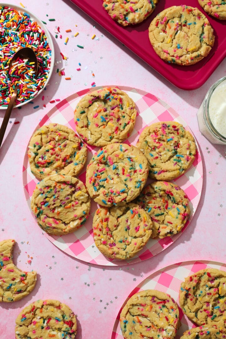 pink gingham print plate with several rainbow sprinkle cookies and a glass of milk off to the right hand side of the photo.