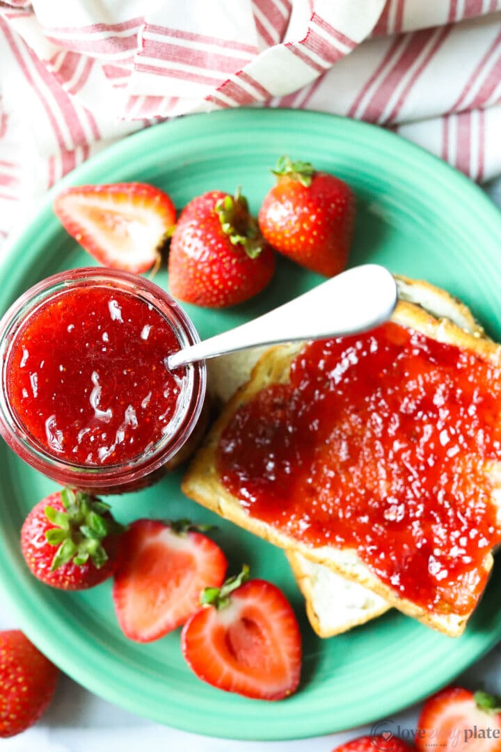 small jar of strawberry jam on a green plate with strawberries and a slice of toast