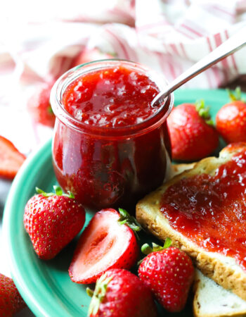 small jar of strawberry jam on a green plate with whole strawberries and a slice of bread