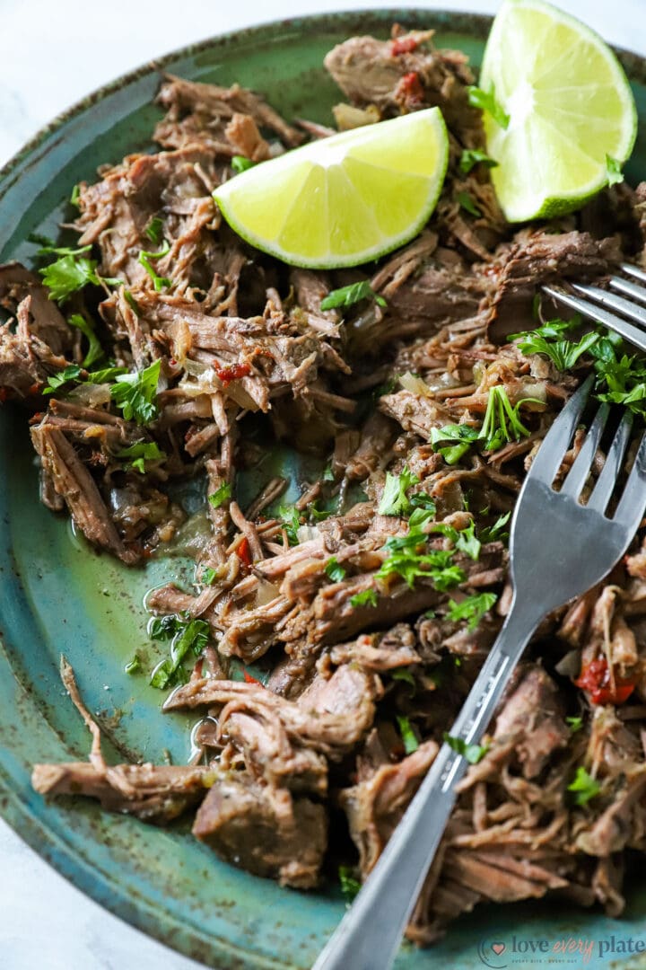 blue bowl of shredded beef with 2 lime wedges and cilantro shreds.
