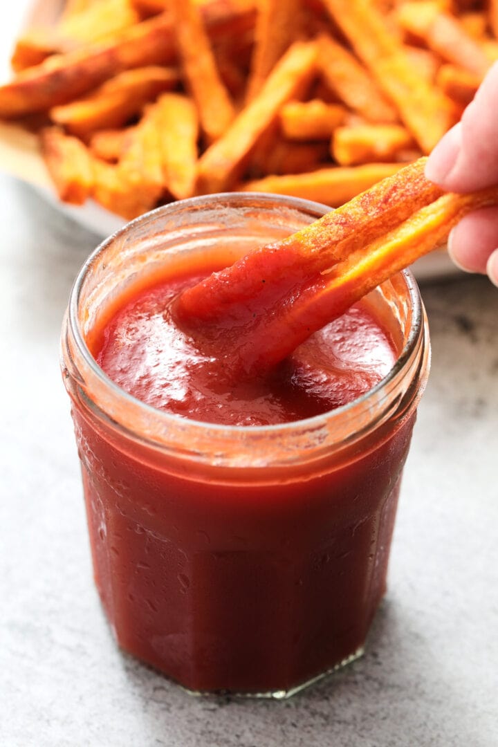 small glass jar of ketchup with a pile of sweet potato fries in the background.