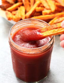 small glass jar of ketchup with a pile of sweet potato fries in the background.