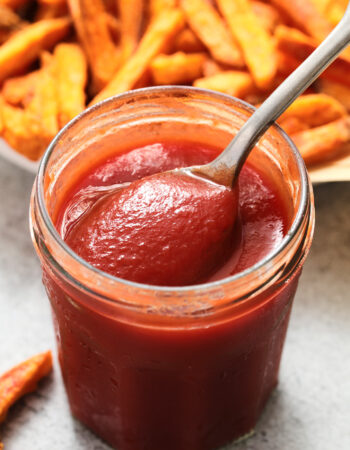 small glass jar of ketchup with a pile of sweet potato fries in the background.