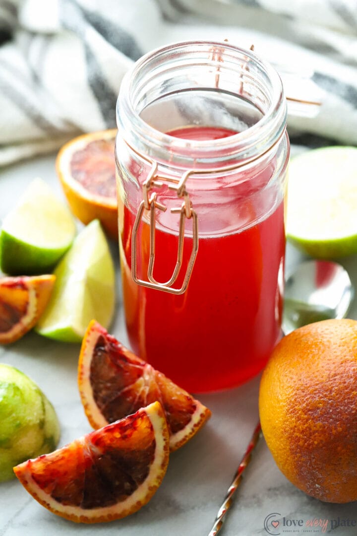 jar of red simple syrup with limes and orange slices