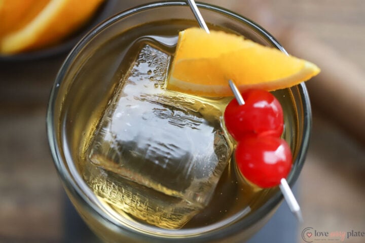 glass holding an old fashioned cocktail garnished with an orange wedge and 2 maraschino cherries
