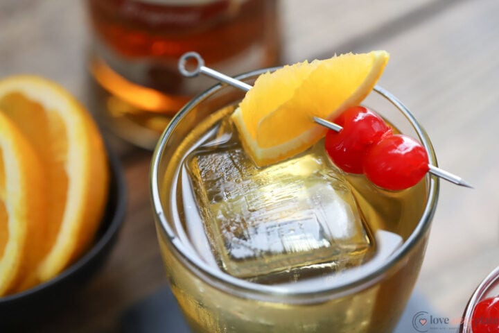 glass holding an old fashioned cocktail garnished with an orange wedge and 2 maraschino cherries