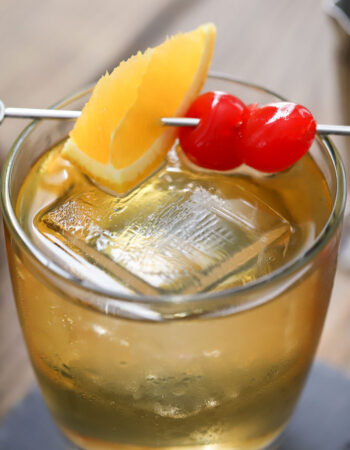 glass holding an old fashioned cocktail garnished with an orange wedge and 2 maraschino cherries