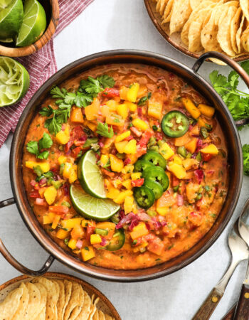 copper bowl of peach mango salsa, garnished with fresh cilantro leaves, lime wedges and sliced jalapenos. There are two bowls of tortilla chips on the right and left sides of the bowl.