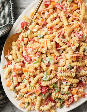 white platter of pasta salad with a gold spoon on the left hand side. There is a green striped towel on the left upper corner.