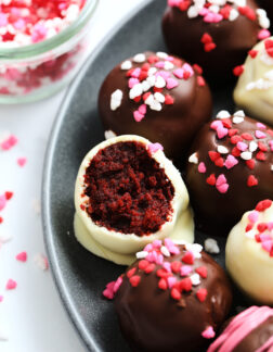 dark plate of red velvet cake balls, covered in valentine's heart sprinkles, one cake bite has been bitten into to reveal the red velvet interior.