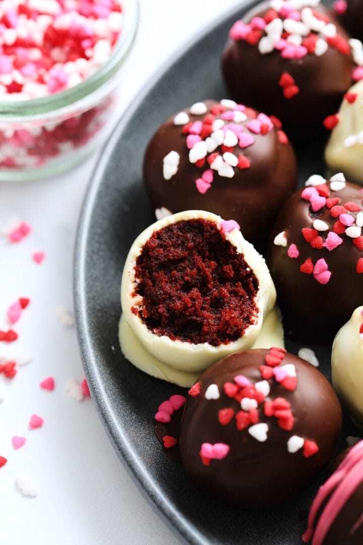 dark plate of red velvet cake balls, covered in valentine's heart sprinkles, one cake bite has been bitten into to reveal the red velvet interior.