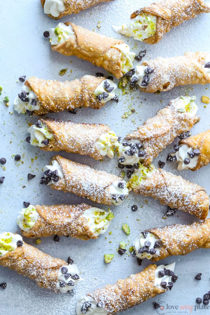 several small cannoli that are garnished with chocolate chips, pistachios and powdered sugar on a gray table.
