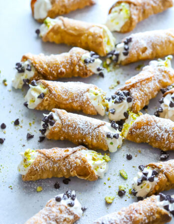 several small cannoli that are garnished with chocolate chips, pistachios and powdered sugar on a gray table.