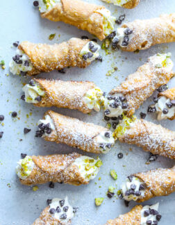 several small cannoli that are garnished with chocolate chips, pistachios and powdered sugar on a gray table.