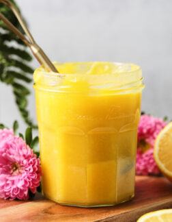small jar of lemon curd with a gold spoon leaning on the left side. There are pink flowers and lemon halves in the background.