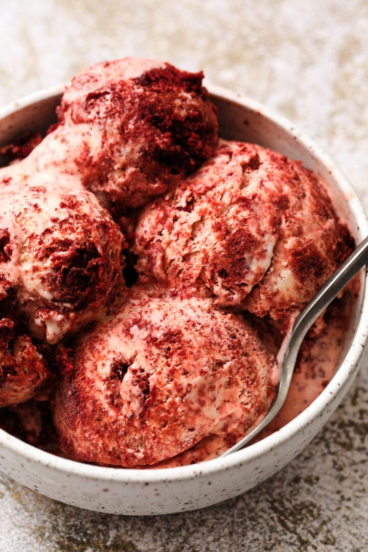 white speckled bowl of red velvet ice cream, with large chunks of red velvet cake and a gray metal spoon on the right side of the bowl.