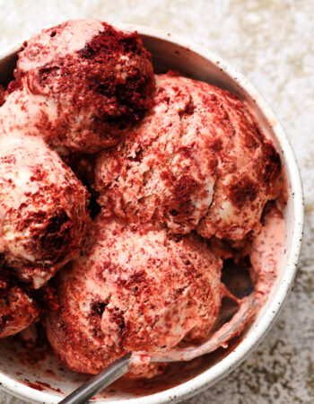 white speckled bowl of red velvet ice cream, with large chunks of red velvet cake and a gray metal spoon on the right side of the bowl.