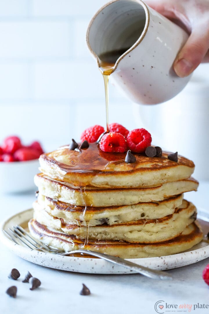 stack of chocolate chip pancakes with raspberries on top, with syrup being poured over top