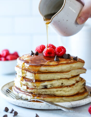 stack of chocolate chip pancakes with raspberries on top, with syrup being poured over top