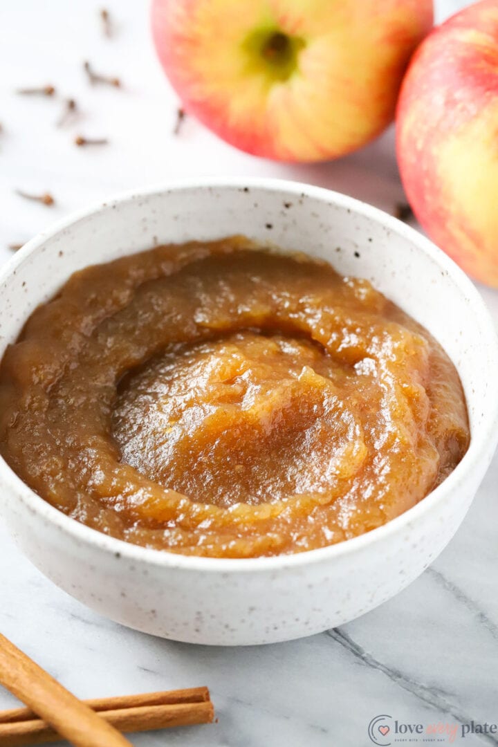 white bowl of apple butter with apples and cinnamon sticks in the background