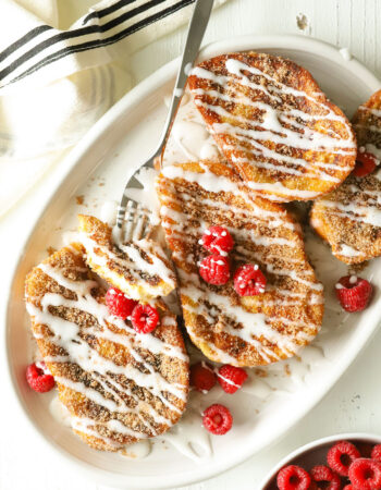 pieces of french toast drizzled with white glaze on a white oval plate with raspberries