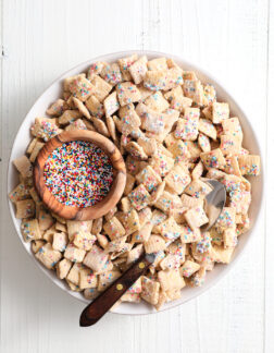 white bowl of rainbow sprinkle cereal snack, with a smaller wooden bowl of rainbow sprinkles off to the left side. There is a wood handled spoon in the bowl.