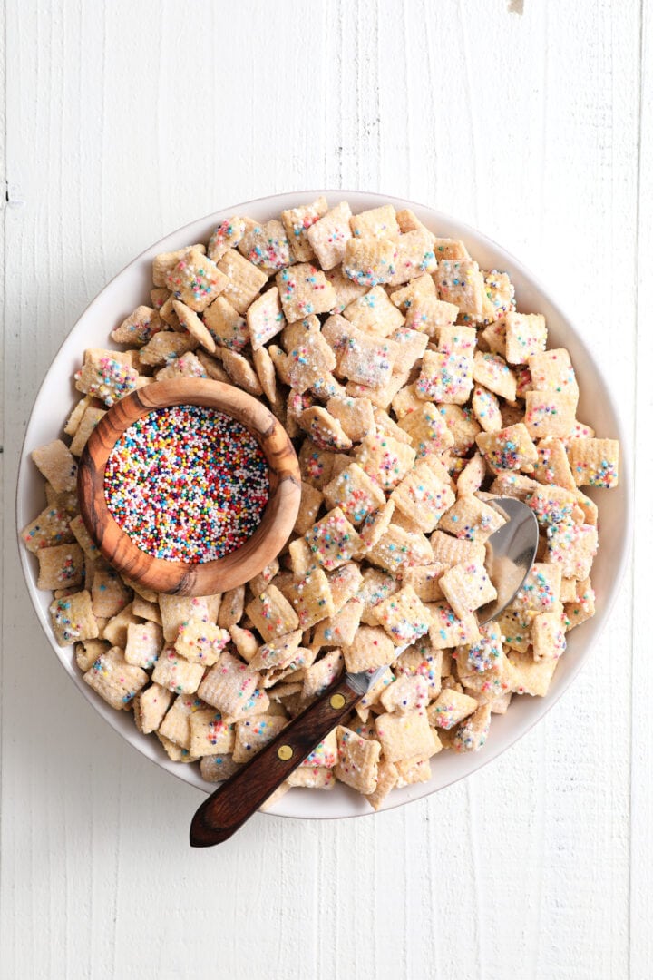 white bowl of rainbow sprinkle cereal snack, with a smaller wooden bowl of rainbow sprinkles off to the left side. There is a wood handled spoon in the bowl.