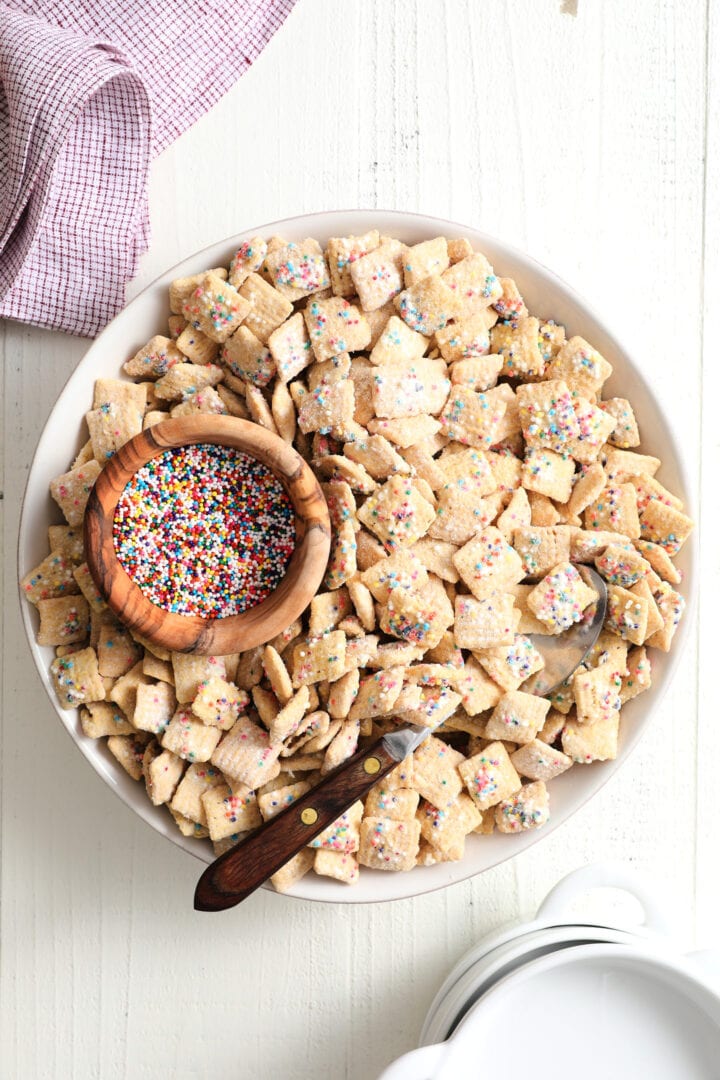 white bowl of rainbow sprinkle cereal snack, with a smaller wooden bowl of rainbow sprinkles off to the left side. There is a wood handled spoon in the bowl.