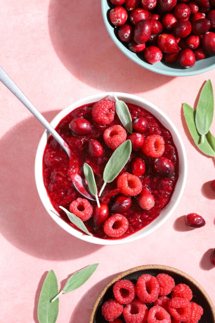 small white bowl of raspberry jam with fresh raspberries, cranberries and sage leaves on top