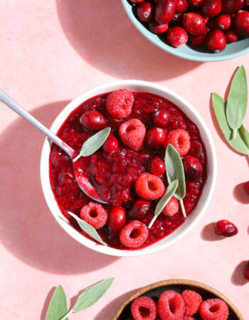 small white bowl of raspberry jam with fresh raspberries, cranberries and sage leaves on top