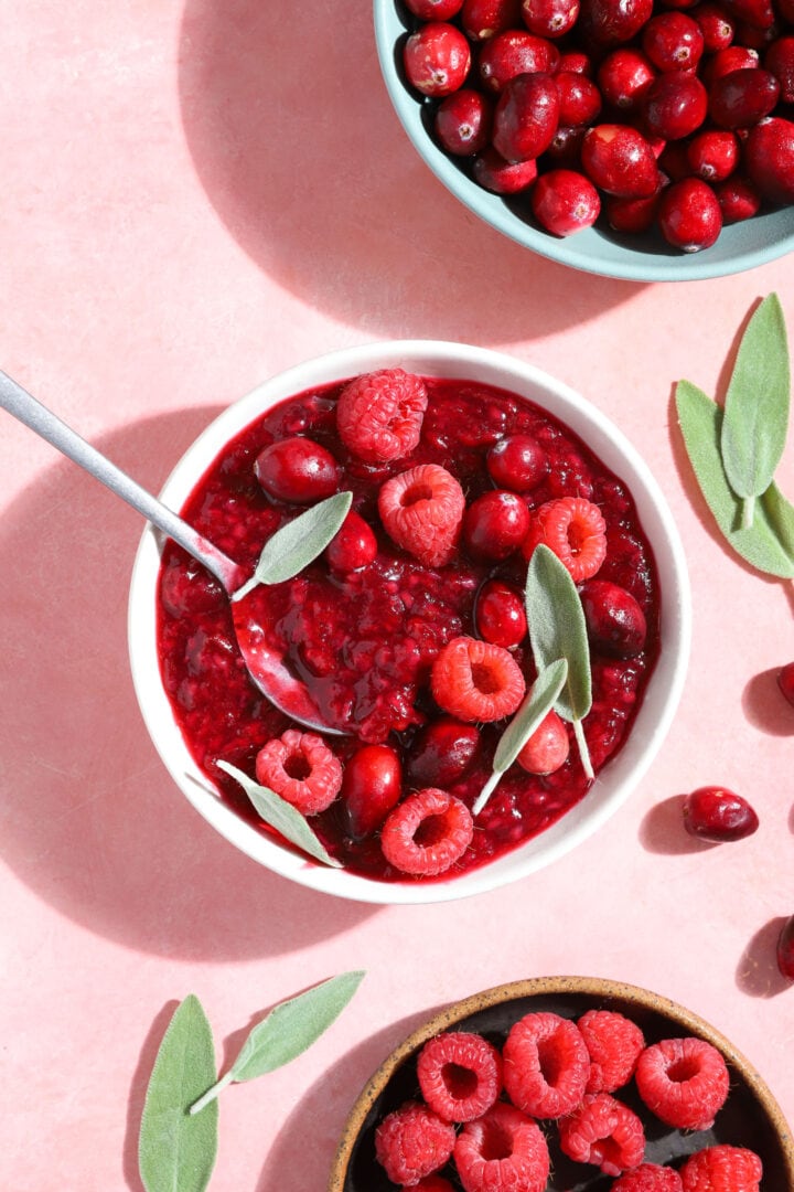 small white bowl of raspberry jam with fresh raspberries, cranberries and sage leaves on top