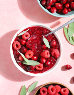 small white bowl of raspberry jam with fresh raspberries, cranberries and sage leaves on top