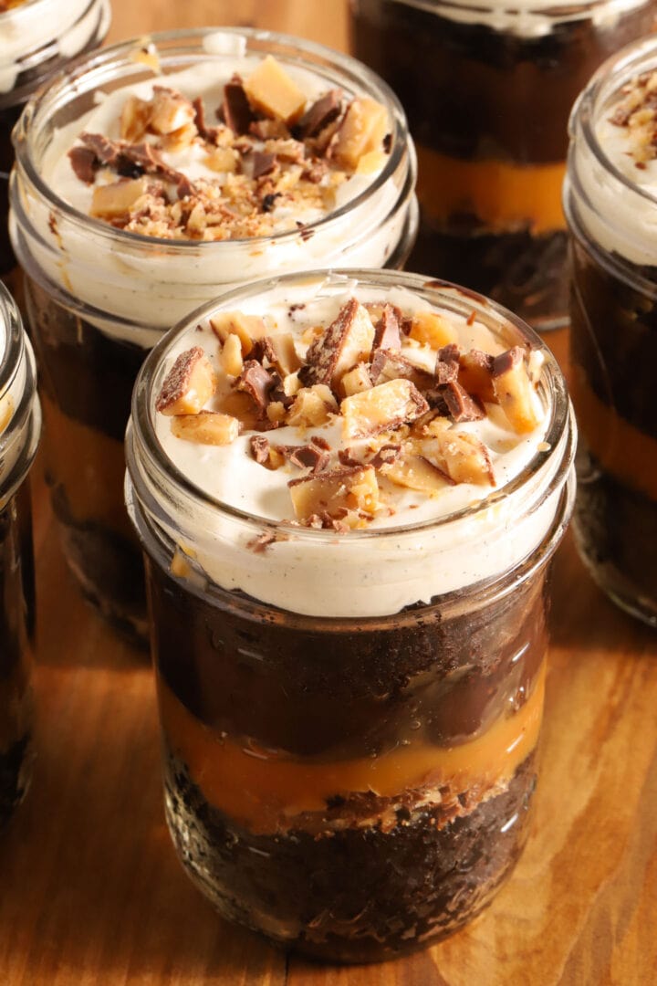 small jar of chocolate cake, chocolate pudding, whipped cream and caramel in layers. A spoon rests on top of the jar with a spoonful of crushed toffee candy bars on top.