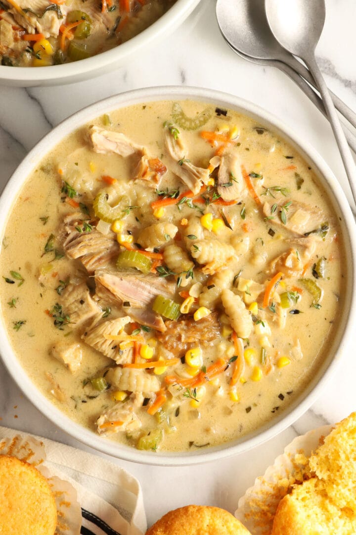 white bowl of creamy turkey soup with vegetables, gnocchi and corn muffins on the side