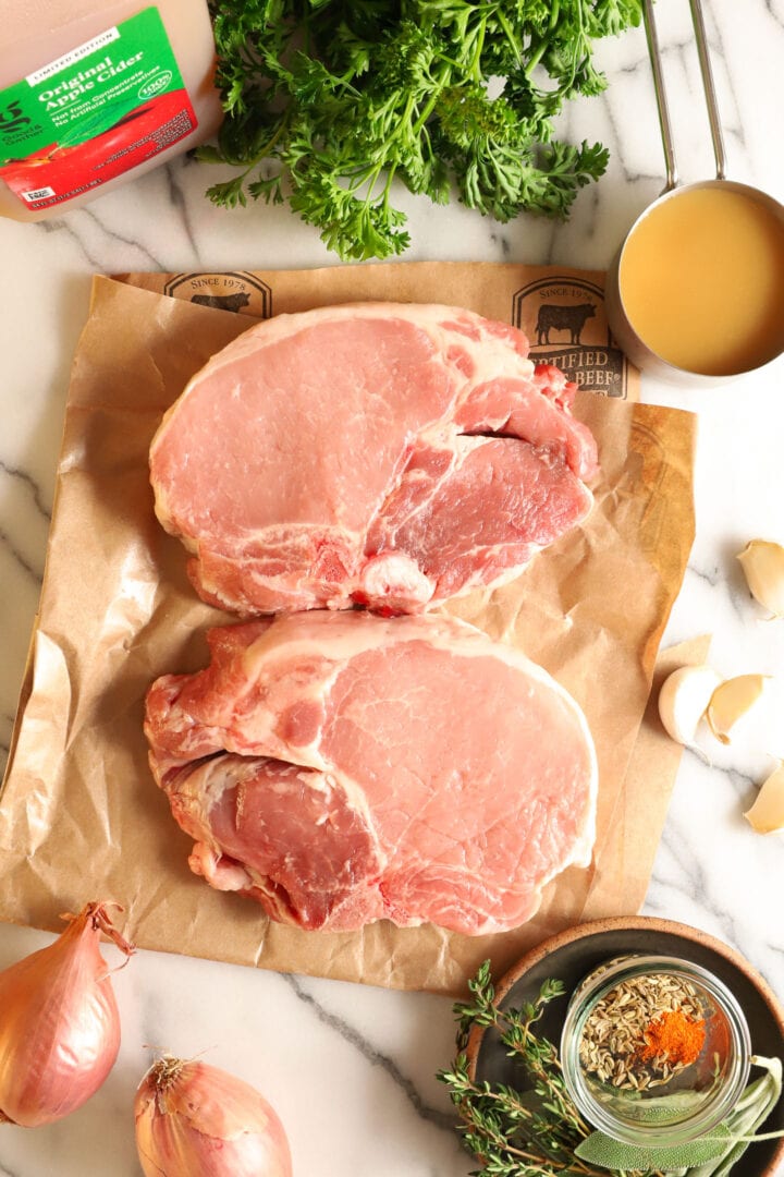 a collection of ingredients to make apple fennel pork chops, including raw pork chops, herbs and spices, and apple cider