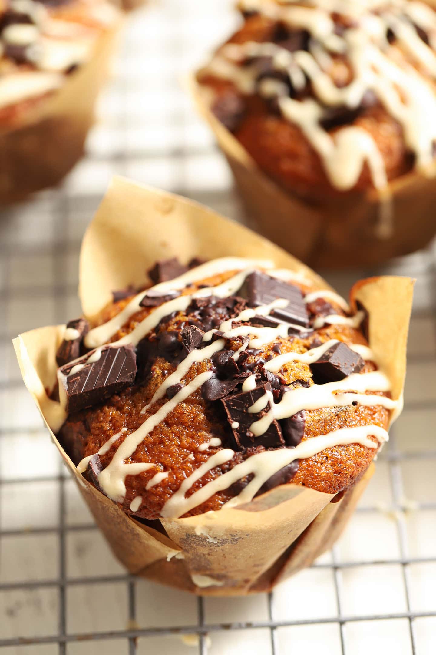 pumpkin chocolate chip muffin with a white glaze drizzled on top, sitting on a cooling rack in a brown paper wrapper.