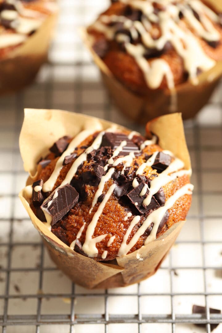 pumpkin chocolate chip muffin with a white glaze drizzled on top, sitting on a cooling rack in a brown paper wrapper.