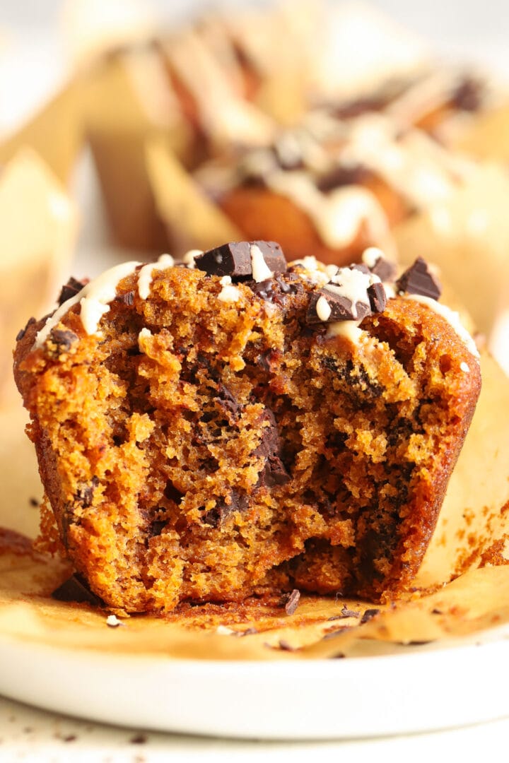 bitten pumpkin chocolate chip muffin with a white glaze drizzled on top, sitting on a white plate in a brown paper wrapper.