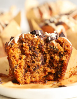 bitten pumpkin chocolate chip muffin with a white glaze drizzled on top, sitting on a white plate in a brown paper wrapper.